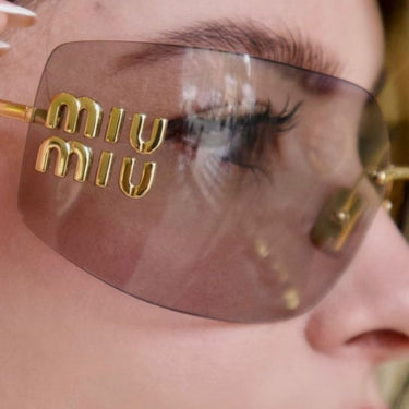 "Gafas MIU MIU con montura metálica dorada, diseño cuadrado y lentes de sol de tonalidad rosa. Destacan los detalles del logo Miu Miu en la parte frontal."